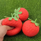 A hand is squeezing a red strawberry-shaped squishy TheMoodease stress ball, alongside two similar strawberry fidget toys on artificial grass, highlighting a fun sensory stress relief toy.